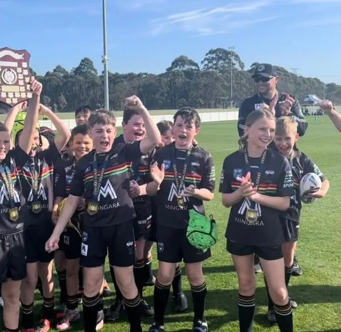 Junior Panthers in black jerseys cheer and clap with their medals on, one holding a premiership shield aloft