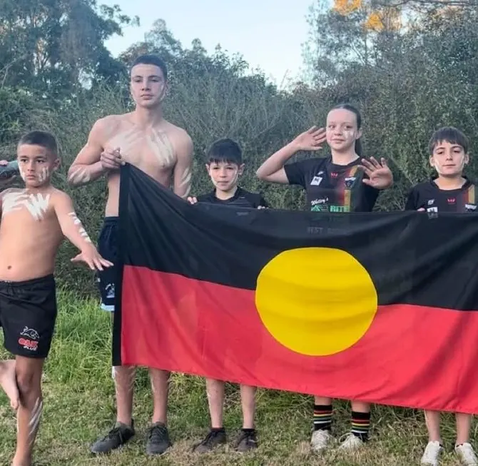 Junior players with traditional ochre markings hold the Aboriginal flag at the edge of the bush