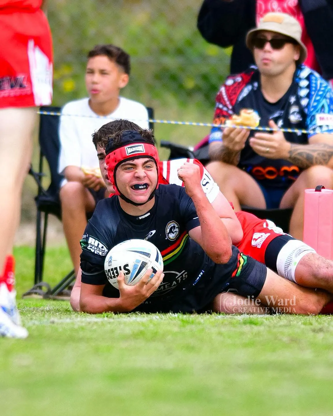 A Panthers junior in red headgear grins and pumps his fist after scoring a try, ball in hand, as spectators watch from the sideline